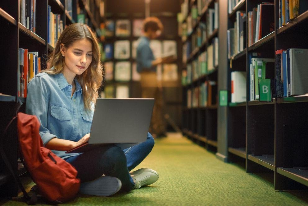 A doctoral student is sitting between two bookshelves on the floor of the library. In her lap is her open computer, in which she is researching the guidelines of good scientific practice.