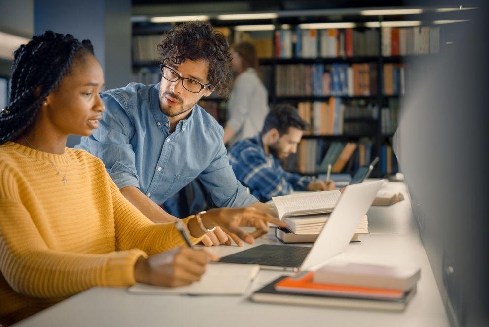 Dekoratives Bild, in dem eine Doktorandin an einem geöffneten Laptop sitzt und eine Frage an den Doktorand neben ihr sitzend bezüglich der Betreungsverereinbarung richtet.