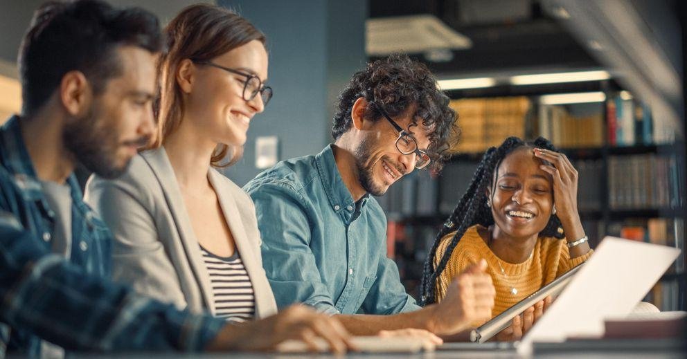 An ethnic and mixed-gender group of young doctoral students sits in the group study room of a library and works together on a project. The mood in the group is good.