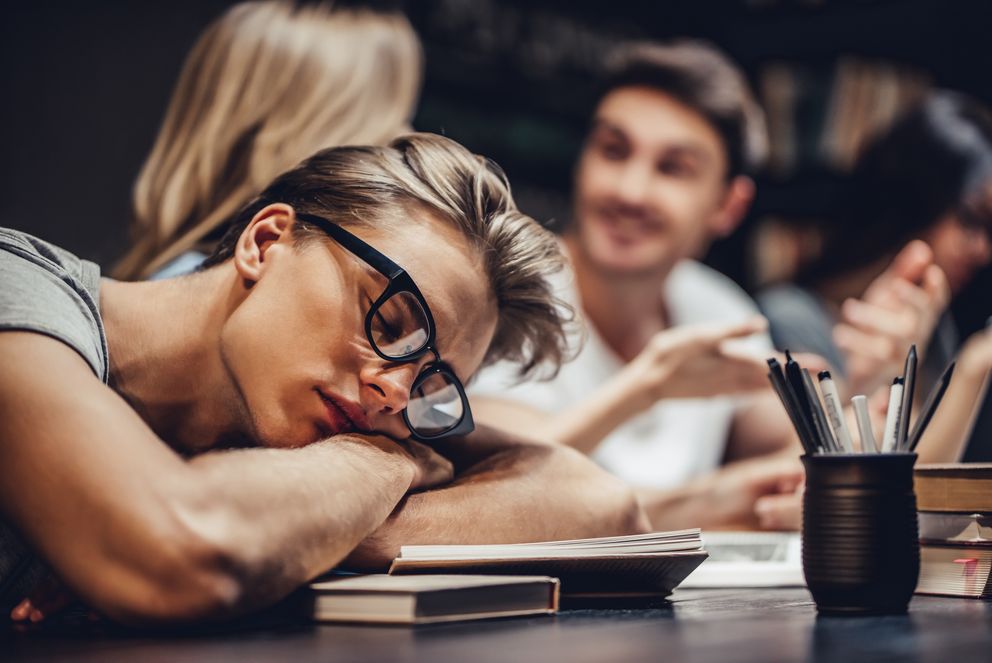 Ein Student mit blondem Haar und Brille ist über seinen Büchern in der Bibliothek eingeschlafen, im Hintergrund sieht man zwei Studierende, die sich unterhalten.