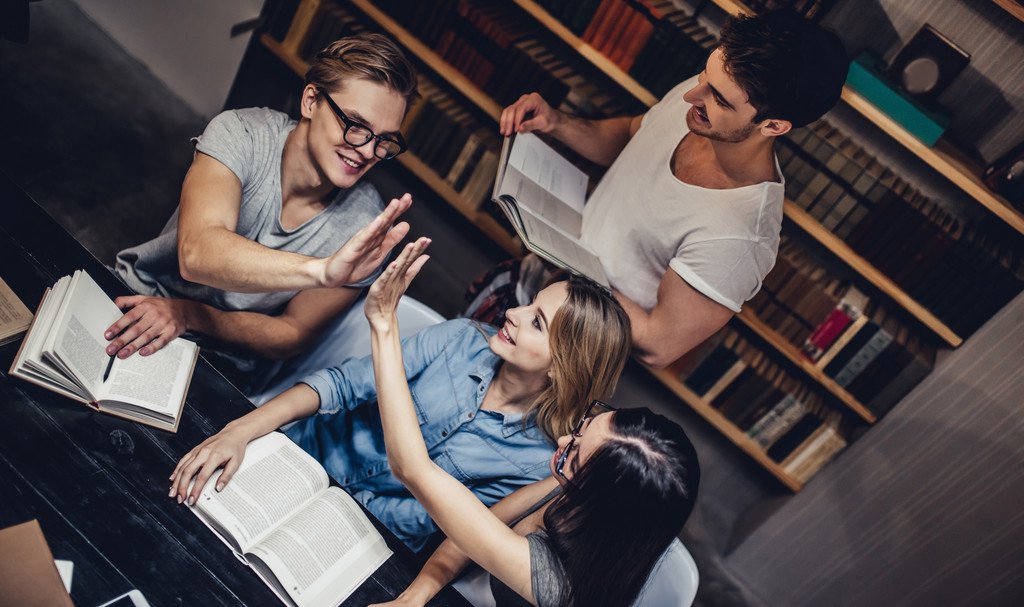 Drei Studierende sitzen über Büchern an einem Tisch in einer Bibliothek, zwei klatschen sich gegenseitig zu. Hinter ihnen steht ein weiterer Student mit einem aufgeschlagenen Buch in der Hand.