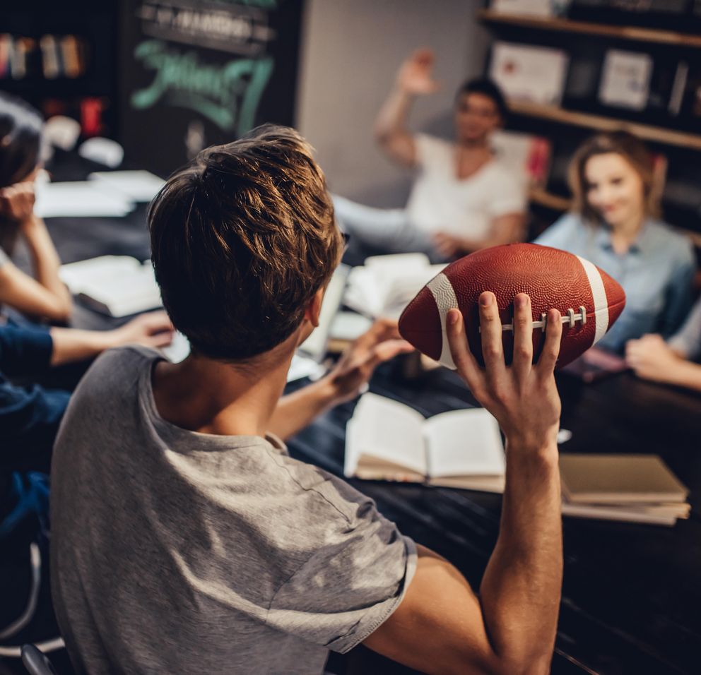 [Translate to Englisch:] Ein Student wirft einem Freund im Hintergrund, der an einem Tisch mit Studierenden sitzt, einen American Football zu.