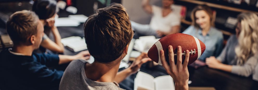 In einer Bibliothek will ein Student einem anderen Studenten, der mit anderen Studierenden an einem Tisch sitzt, einen American Football zuwerfen.