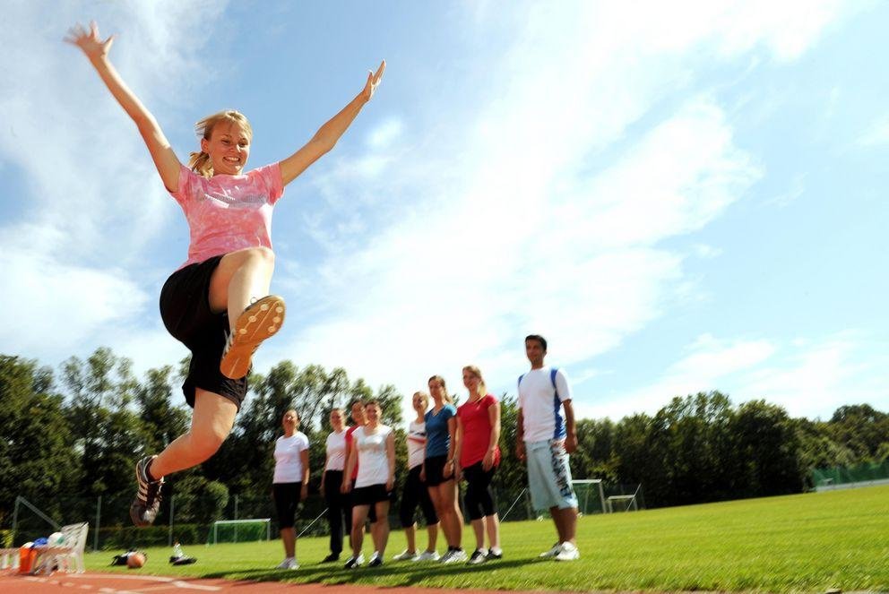 Promotionsinteressierte Das Bild zeigt eine Gruppe von Studierenden auf dem Sportplatz. Eine Studentin setzt zum Weitsprung an.