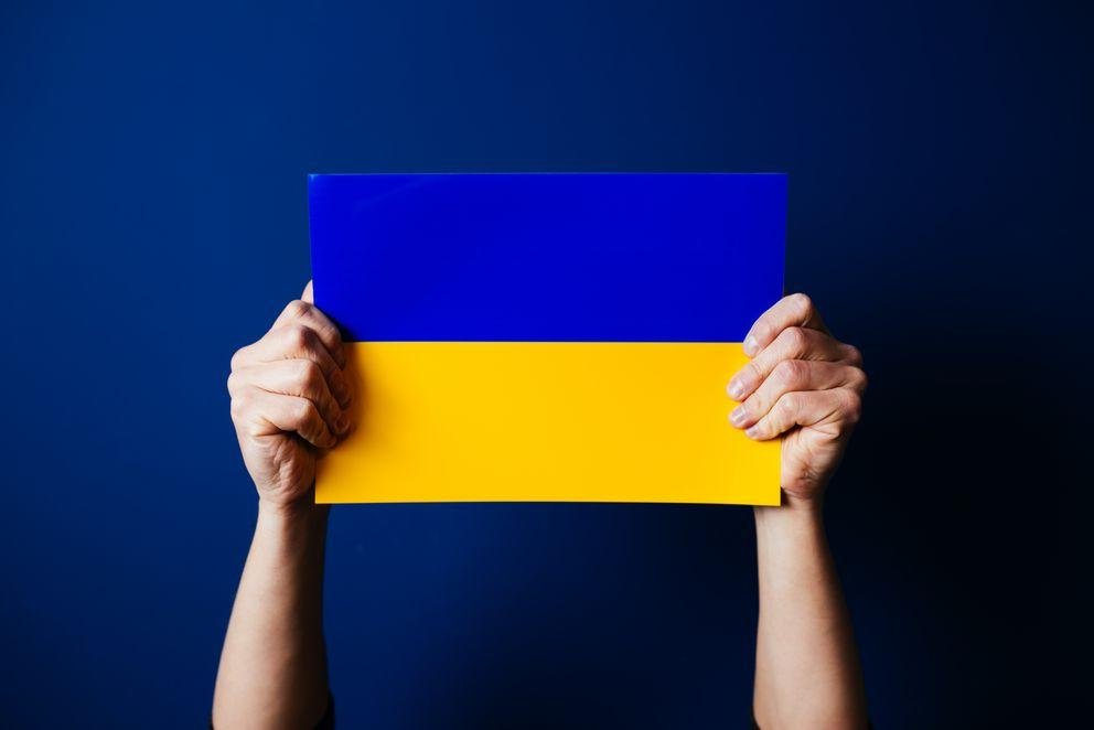 Zwei Hände halten eine ukrainische Flagge vor blauem Hintergrund hoch.