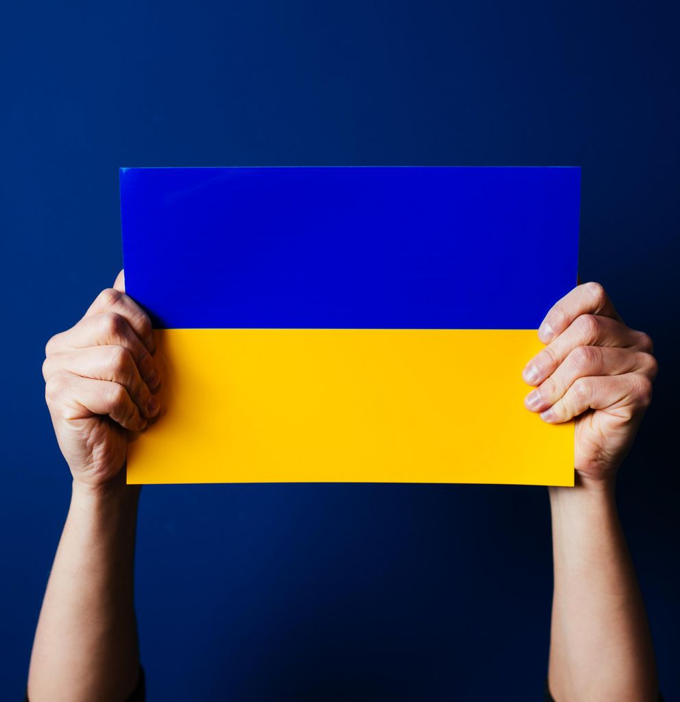 Zwei Hände halten eine ukrainische Flagge vor blauem Hintergrund hoch.