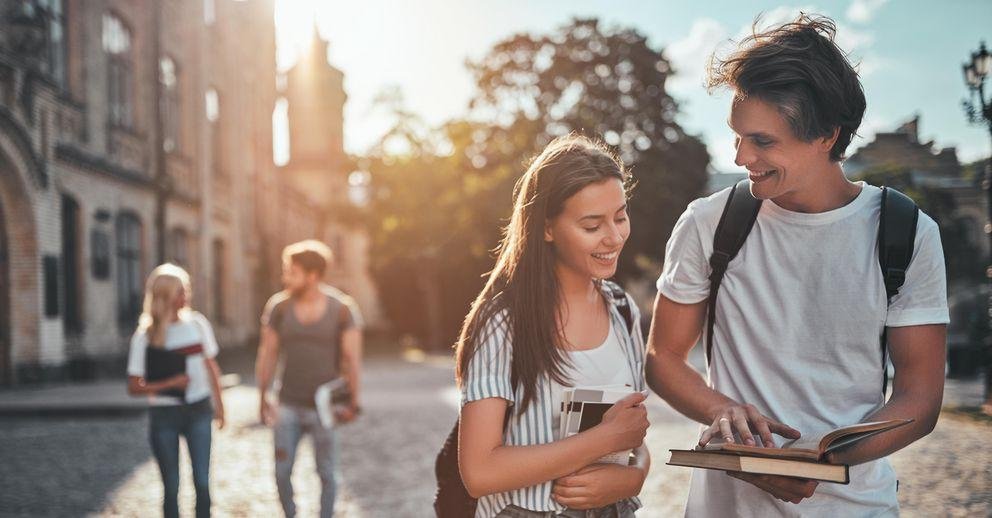 [Translate to Englisch:] Ein Student zeigt im Vordergrund einer Studentin etwas in einem Buch. Im Hintergrund sieht man ein Universitätsgebäude und zwei sich miteinander unterhaltende Studierende.