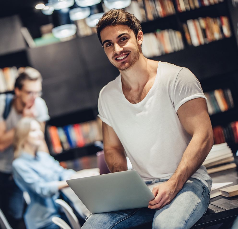 Ein Student sitzt mit einem Laptop auf dem Schoß auf einem Tisch in einer Bibliothek, im Hintergrund sieht man eine Studentin, die Bücher trägt, und einen Studierenden sowie eine Studierende, die gemeinsam auf einen Laptop schauen.