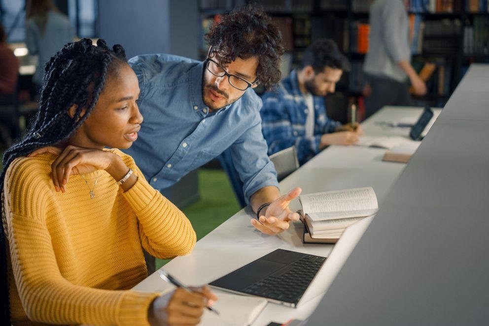 Dekoratives Bild zur Auflockerung des Textes zum Wissenschaftszeitvertragsgesetz, das eine Promovierende zeigt, die grübelnd über ihrem Laptop sitzt. Neben ihr sitzt ein anderer Promovierender der ihr das Wissenschaftszeitvertragsgesetz erklärt.