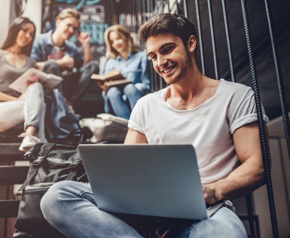 Ein Student mit weißem T-Shirt sitzt auf einer Treppe mit seinem Laptop auf dem Schoß, über ihm sitzen drei weitere Studierende.