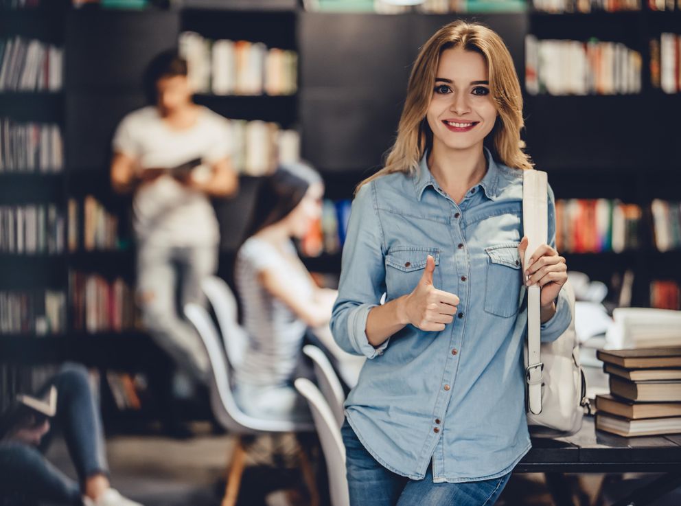 [Translate to Englisch:] Ein junge Frau mit einer weißen Tasche über der Schulter steht lächelnd in einer Bibliothek mit Studierenden im Hintergrund und zeigt der Betrachterin bzw. dem Betrachter ihren Daumen.