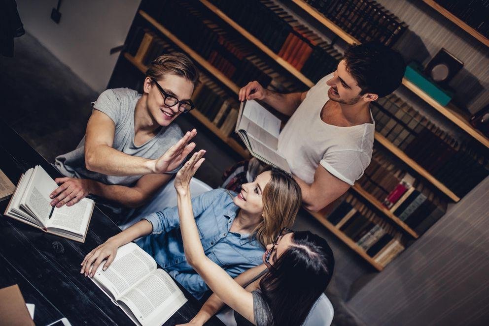 [Translate to Englisch:] Drei Studierende sitzen über Büchern an einem Tisch in einer Bibliothek, zwei klatschen sich gegenseitig zu. Hinter ihnen steht ein weiterer Student mit einem aufgeschlagenen Buch in der Hand.