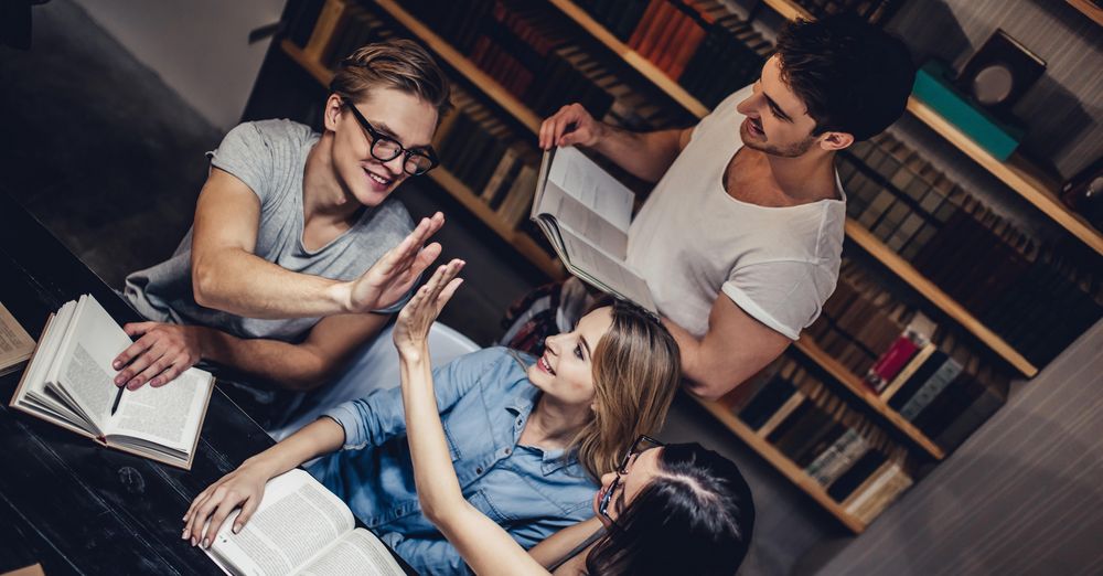 Drei Studierende sitzen über Büchern an einem Tisch in einer Bibliothek, zwei klatschen sich gegenseitig zu. Hinter ihnen steht ein weiterer Student mit einem aufgeschlagenen Buch in der Hand.