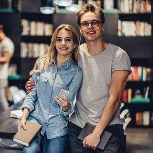 Eine Studentin und ein Student stehen in einer Bibliothek. Er umarmt sie, sie hält ein Handy in der Hand und beide teilen sich die dazugehörigen Kopfhörer.