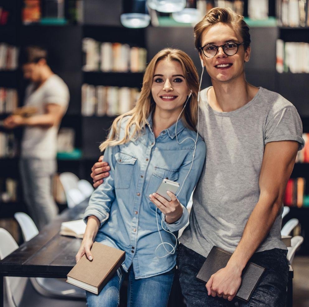 Eine Studentin und ein Student stehen in einer Bibliothek. Er umarmt sie, sie hält ein Handy in der Hand und beide teilen sich die dazugehörigen Kopfhörer.