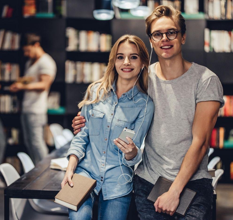 Eine Studentin und ein Student stehen in einer Bibliothek. Er umarmt sie, sie hält ein Handy in der Hand und beide teilen sich die dazugehörigen Kopfhörer.